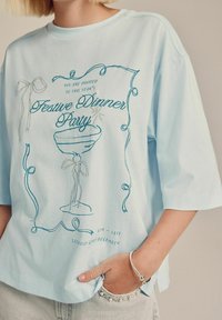 Person, die ein hellblaues T-Shirt mit einem festlichen Einladungdesign für ein Abendessen und helle Denim-Jeans trägt, mit der Hand in der Tasche und einem silbernen Armband.