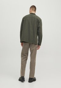 Uomo in piedi di spalle, con una giacca verde scuro, pantaloni beige, scarpe nere e un orecchino di cerchio argentato su uno sfondo chiaro e semplice.