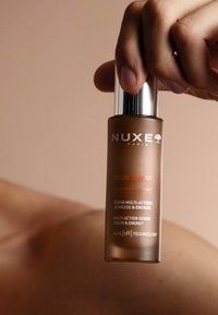 Brązowa szklana butelka na serum z srebrną nakrętką, z białym i pomarańczowym tekstem marki oraz szczegółami produktu. Gładka tekstura, cylindryczny kształt.