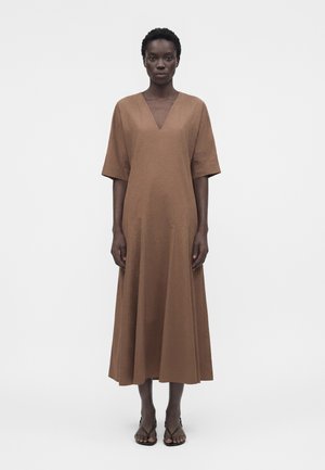 Mujer de pie mirando hacia adelante, con un vestido midi marrón de manga corta y escote en V, combinado con sandalias negras de punta abierta sobre fondo blanco.