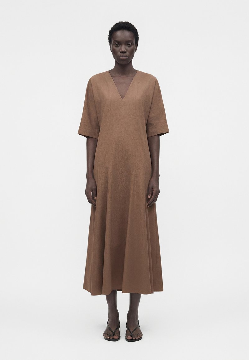 Γυναίκα που στέκεται με το πρόσωπο προς τα εμπρός, φοράει ένα καφέ midi φόρεμα με κοντά μανίκια και V-λαιμόκοψη, συνδυασμένο με μαύρα σανδάλια ανοιχτής μύτης σε λευκό φόντο.