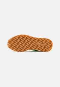 Semelle de chaussure verte avec une surface en caoutchouc orange texturée et ondulée. Présente un détail de marque moulé et des bords arrondis.