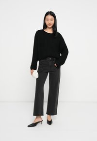 Maglione nero a maglia grossa, jeans neri a vita alta, mule nere con tacco, borsa bianca a pieghe. Un outfit semplice e rilassato su uno sfondo neutro.