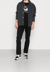 Camicia a righe nera e grigia con bottoni, t-shirt grafica nera sotto, pantaloni neri e sneakers bianco e neri.