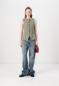 Oliven grønn ermeløs vest med fem knapper, kombinert med brede lyseblå jeans. Holder en liten rød håndveske. Svart fottøy.