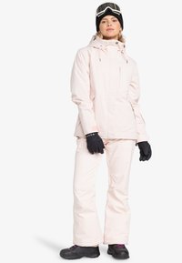 Roze sneeuwjack met verstelbare capuchon, voorzakken en elastische manchetten, gecombineerd met bijpassende lichtroze sneeuwbroek en zwarte handschoenen.