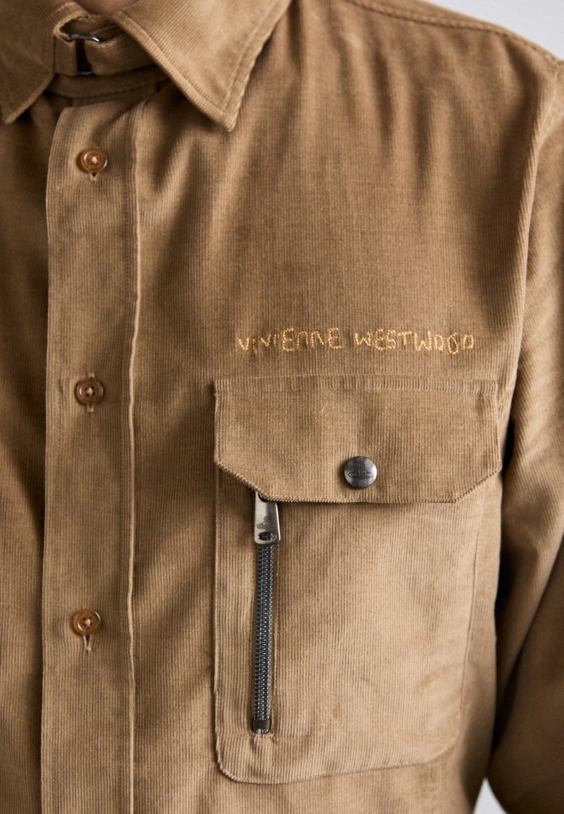 Chemise en velours côtelé marron avec un boutonnière à l'avant, un logo brodé et une poche poitrine avec fermeture éclair. Tissu texturé avec des côtes verticales.