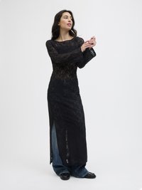 Vestido de renda preta com um corpete ajustado, mangas longas e largas, e aberturas laterais. Abaixo, jeans de ganga azuis largos e sapatos pretos completam o visual.