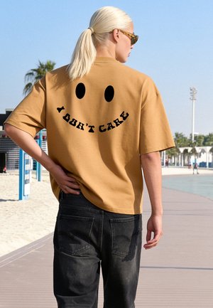Personne aux cheveux clairs attachés en queue de cheval porte un t-shirt beige avec un visage souriant noir et le texte "I DON'T CARE", marchant sur une promenade de plage ensoleillée.