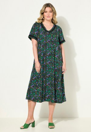 Zwart bloemenprintjurk met groene, paarse en gele patronen. Losse pasvorm, korte mouwen en een V-halslijn. Gecombineerd met groene blokhak sandalen.
