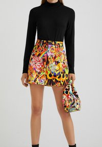 Donna con un top nero a maniche lunghe e una mini gonna colorata con motivi di fiamme, fiori e creature mitiche, mentre tiene in mano una borsa a tracolla coordinata.