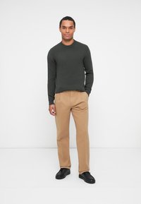Pull en tricot vert foncé, pantalon beige et baskets noires. Le modèle se tient debout contre un fond blanc avec un éclairage uniforme.