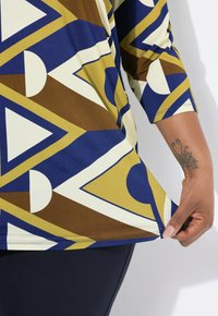 Mehrfarbige Tunika mit geometrischen Formen in Blau, Gelb, Weiß und Braun; glattes Material mit Dreiviertelärmeln und einem abgerundeten Saum.