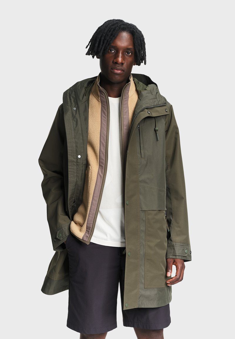 Homme aux cheveux bruns tressés portant une veste olive en couches, un gilet en polaire beige, une chemise blanche et un short noir, debout devant un fond blanc.