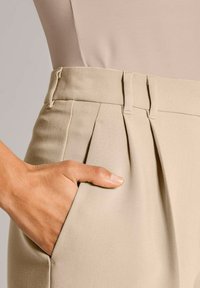 Hand met een bruine huid rustend in de zak van beige high-waisted pants, draagt een lichtbeige passend bovenstuk.