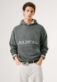 Felpa grigia con cappuccio e logo frontale "PJL1973Club", tessuto testurizzato e polsini sovrapposti su pantaloni bianchi. Design minimalista e vestibilità casual.