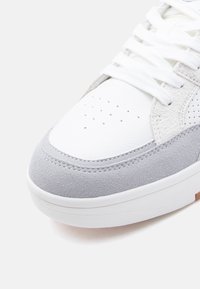 Κοντινό πλάνο άκρου λευκού αθλητικού παπουτσιού με διάτρητο δέρμα, γκρίζες επενδύσεις από σουέτ, λευκά κορδόνια και λευκή λαστιχένια σόλα.