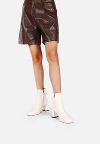 Stivaletti bianchi alla caviglia con punta quadrata e tacco spesso, dotati di zip laterale e una texture liscia e lucida. Abbinati a pantaloni corti color marrone cioccolato.