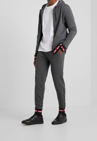 Grauer Reißverschluss-Hoodie mit kontrastierenden roten und schwarzen Akzenten, kombiniert mit grauen Jogginghosen. Schwarze Sneakers und schwarze Socken mit sichtbaren Logo-Details.