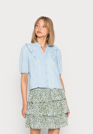 Blusa de botão azul clara com decote franzido e mangas bufantes, combinada com uma saia verde, em camadas, com padrão.