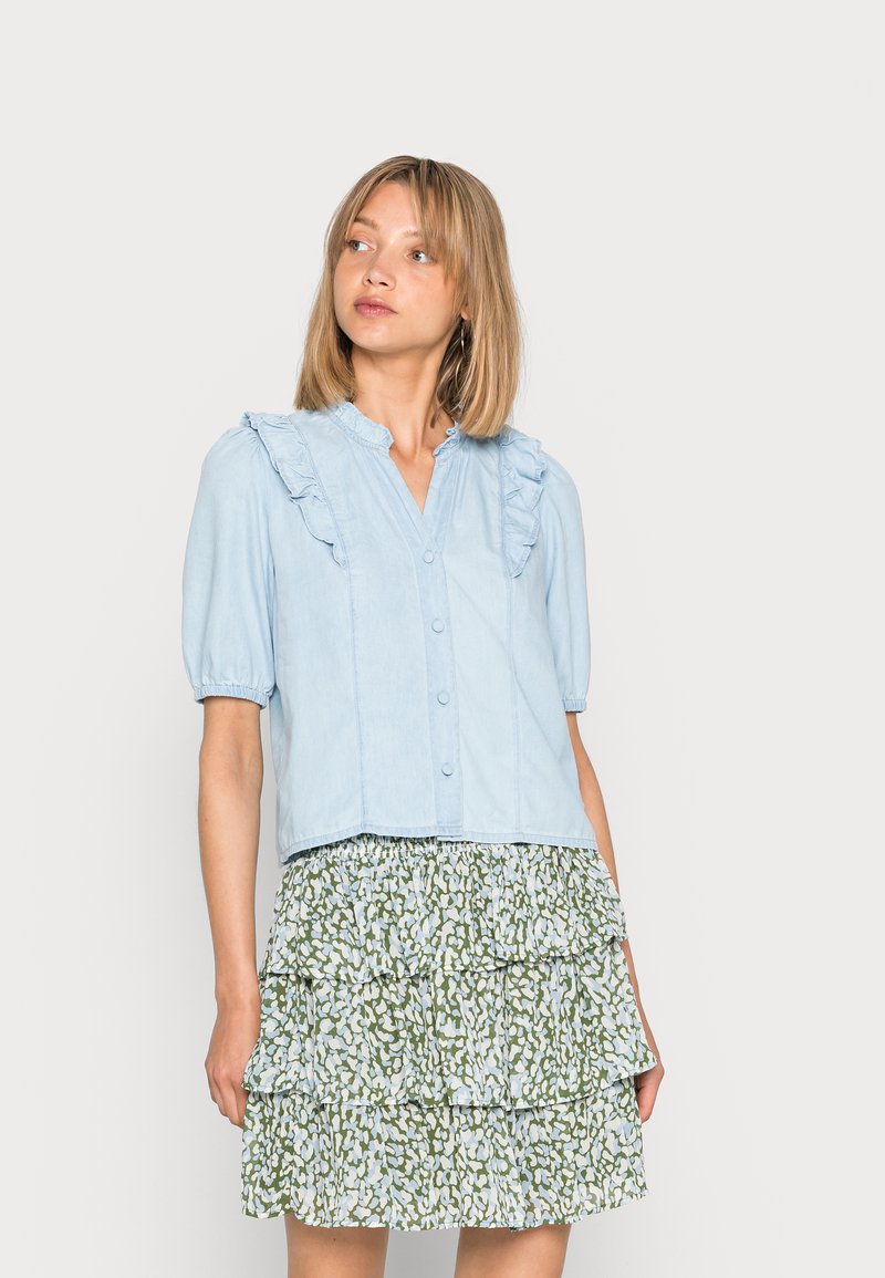 Blusa de botão azul clara com decote franzido e mangas bufantes, combinada com uma saia verde, em camadas, com padrão.