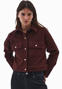 Burgundowa kurtka z cordurowego materiału z kołnierzem, dwoma kieszeniami na piersi, złotymi guzikami i skróconym fasonem. Posiada widoczną teksturę i dopasowany krój.