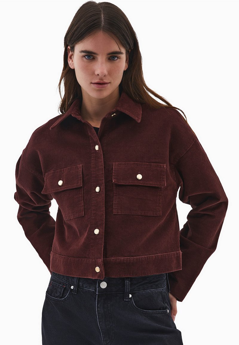 Burgundowa kurtka z cordurowego materiału z kołnierzem, dwoma kieszeniami na piersi, złotymi guzikami i skróconym fasonem. Posiada widoczną teksturę i dopasowany krój.