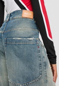 Osoba wearing vyblednuté modré džínsy s koženou nášivkou značky Replay a čiernym, červeným a bielym topom s dlhým rukávom.