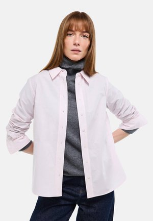 Femme aux cheveux roux lisses portant une chemise rose clair boutonnée sur un pull gris à col roulé et un pantalon foncé, debout les mains sur les hanches.