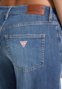 Mėlyni džinsai su rudos odos etikete „GUESS JEANS“ ant juosmens, su trikampiu logotipu ant nugarinės kišenės.