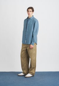 Svetlo modra srajca s kratkimi rokavi in sproščenim krojem, v kombinaciji s svetlo rjavimi širokimi hlačami in belimi športnimi čevlji. Gladek material z minimalnimi detajli.