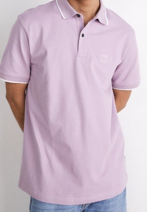 Fioletowa koszulka polo z białym obszyciem na kołnierzyku i rękawach, z dwoma guzikami oraz delikatnym logo marki na piersi.