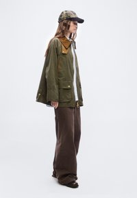 Γυναίκα φοράει ένα φαρδύ παλτό σε ελαιί πράσινο χρώμα με καφέ γιακά, φαρδιά καφέ παντελόνια, λευκό πουκάμισο, σκούρα παπούτσια και καρό καπέλο, στραμμένη στο πλάι.