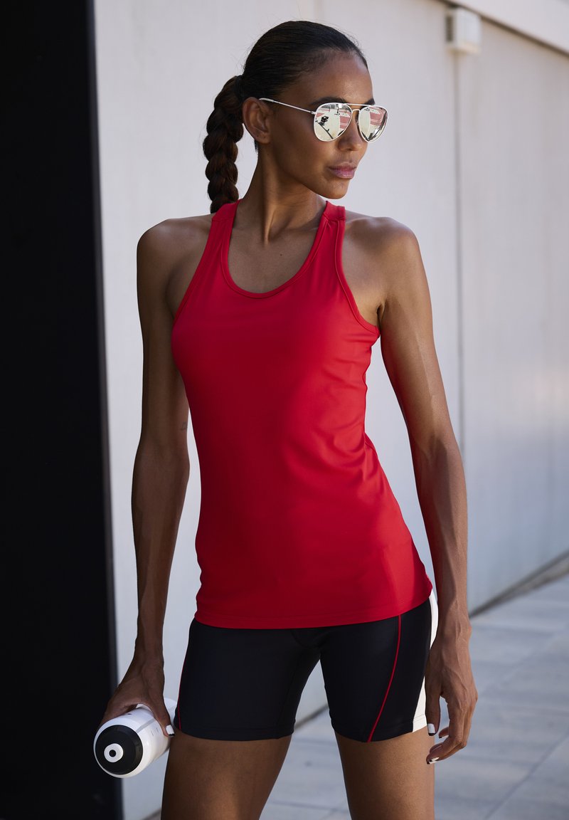 Rotes sportliches Tanktop mit Rundhalsausschnitt, kombiniert mit schwarzen Shorts mit roten und weißen Akzenten. Hält eine weiße Sportwasserflasche.