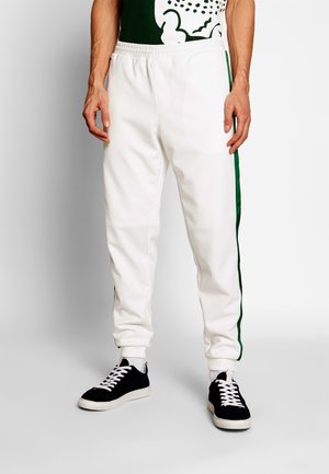 Homme portant un pantalon de jogging blanc avec des bandes vertes sur les côtés, des baskets noires avec des lacets blancs et un haut graphique vert foncé, debout sur un sol clair.