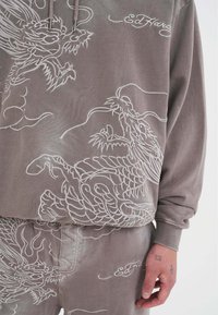 Felpa grigia con ricamo di drago bianco e logo. Presenta una vestibilità comoda, cappuccio con cordino e pantaloni abbinati. La texture sembra morbida.