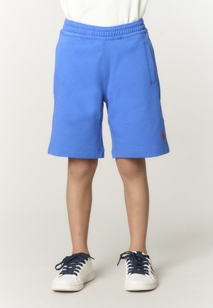 Bambino che indossa pantaloncini di cotone blu con vita elastica e sneakers bianche con lacci blu scuro, in piedi davanti a uno sfondo chiaro e uniforme.