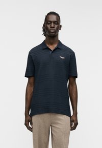 Μπλε ναυτικό πόλο μπλουζάκι με οριζόντιες ρίγες, κοντά μανίκια και γιακά. Διαθέτει μικρό μεταλλικό λογότυπο στο στήθος και άνετη εφαρμογή.