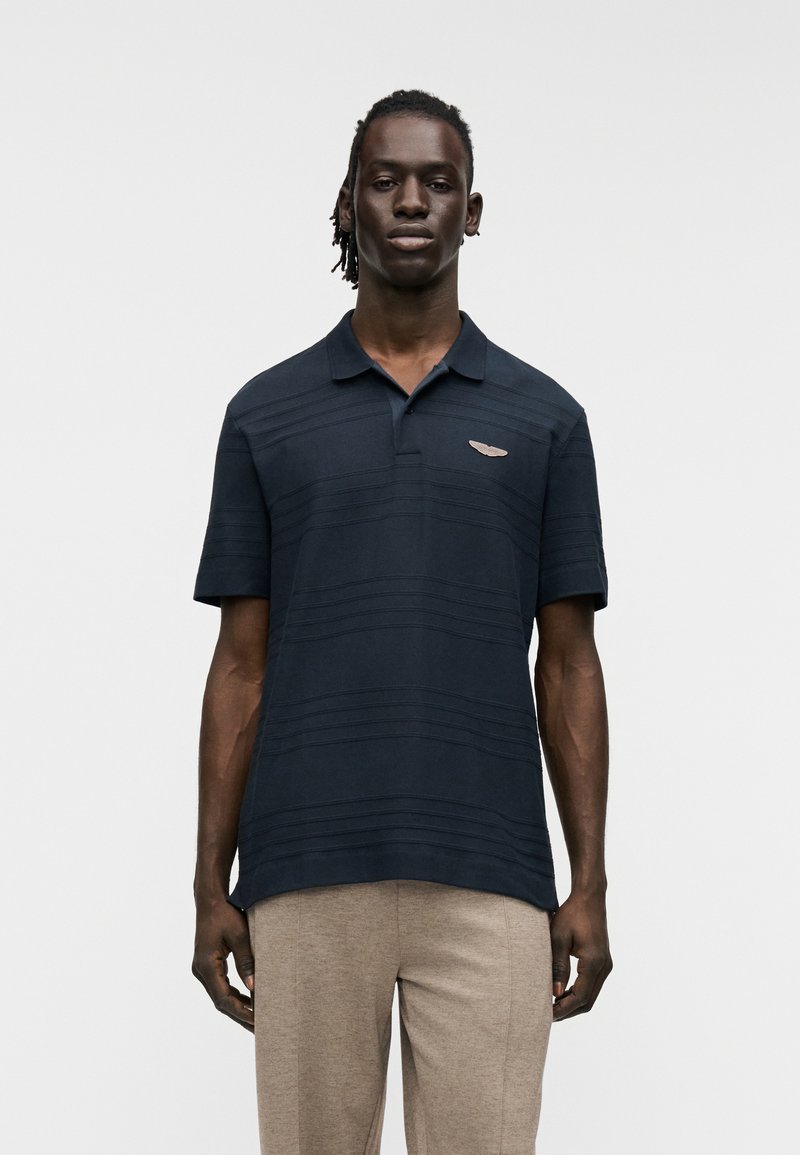 Μπλε ναυτικό πόλο μπλουζάκι με οριζόντιες ρίγες, κοντά μανίκια και γιακά. Διαθέτει μικρό μεταλλικό λογότυπο στο στήθος και άνετη εφαρμογή.