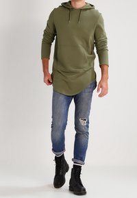 Sudadera verde oliva con mangas largas y bolsillo delantero, combinada con unos vaqueros desgastados azul y botas negras de cordones.