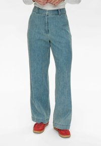 Világoskék, bő szárú farmernadrág sima textúrával, magas derékkal és elöl redős részletekkel. Piros sportcipővel párosítva, barna talppal.