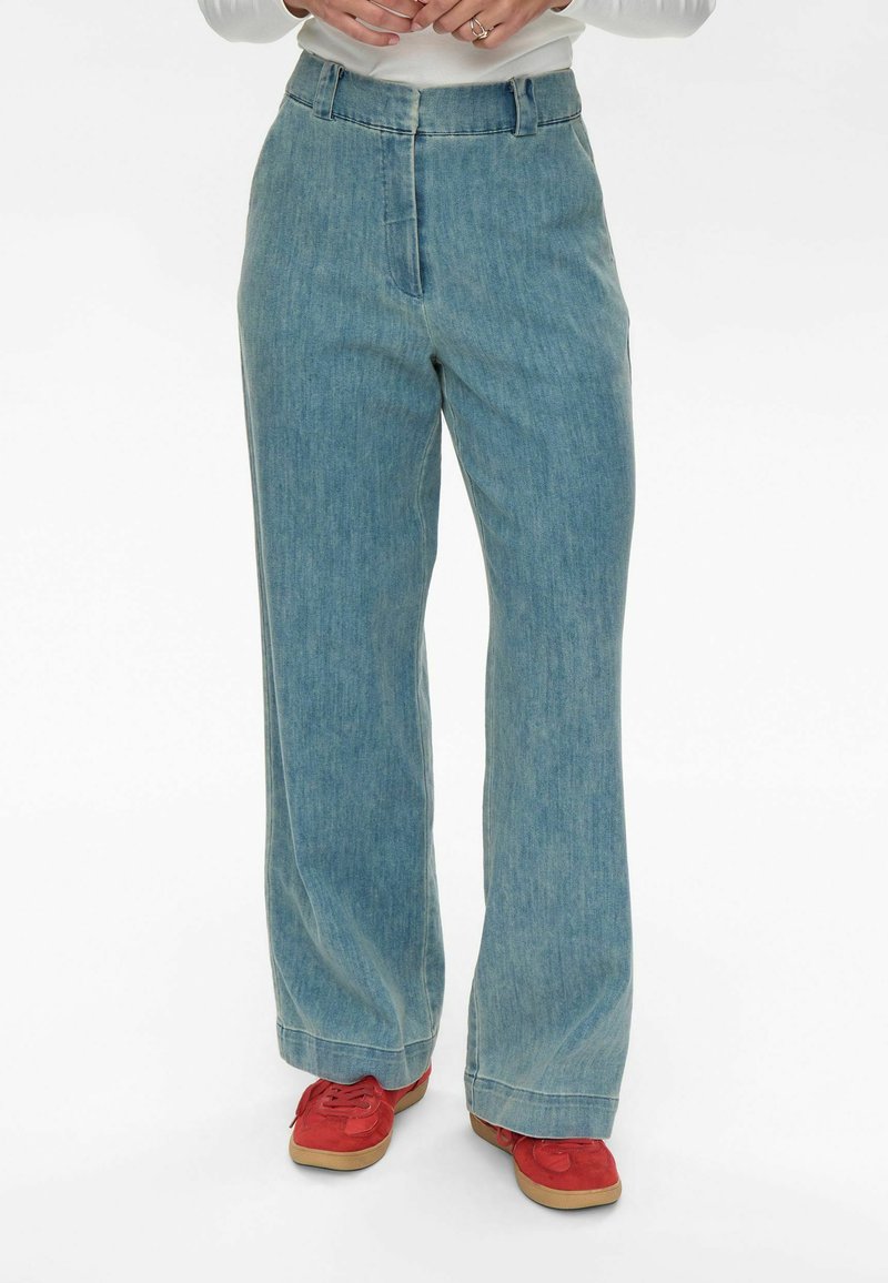 Világoskék, bő szárú farmernadrág sima textúrával, magas derékkal és elöl redős részletekkel. Piros sportcipővel párosítva, barna talppal.