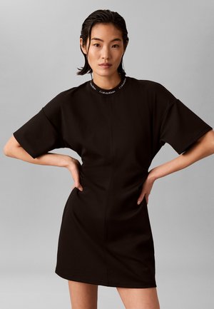 Giovane donna con capelli effetto bagnato che indossa un vestito nero Calvin Klein a maniche corte, mani sui fianchi, sfondo grigio neutro.