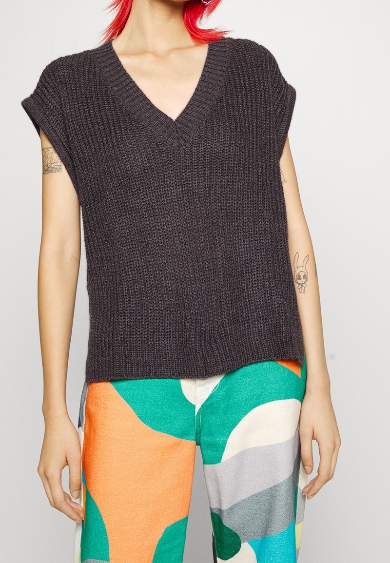 Personne aux cheveux roux portant un haut sans manches en maille gris foncé et un pantalon coloré à motifs abstraits verts, orange et gris.