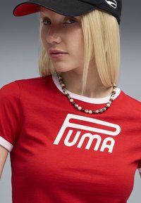 Röd bomullströja med vit kant och stor "PUMA"-logotyp, i kombination med ett pärlhalsband och en svart keps med logotyp.