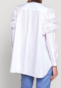 Witte blouse met getextureerde, gerimpelde mouwdetails. De voorkant heeft een klassieke kraag, de achterkant is iets langer met een ronde zoom.