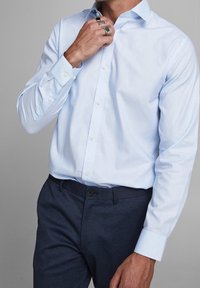 Jasnoniebieska koszula z gładkiego materiału z długimi rękawami i zapiętymi mankietami, w zestawieniu z ciemnymi spodniami w wzory. Zbliżenie na regulację kołnierza.
