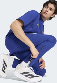 Niebieska sportowa koszulka z krótkim rękawem i graficznym wzorem, w połączeniu z niebieskimi zwężanymi spodniami oraz białymi butami z czarnymi paskami.