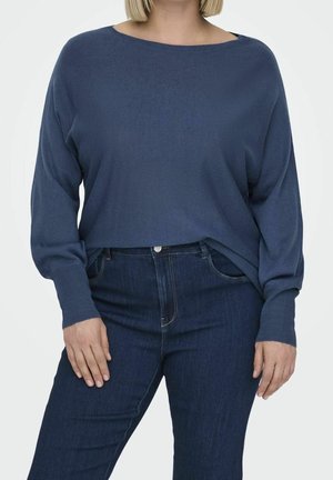Personne portant un pull bleu marine ample et un jean bleu foncé taille haute, debout avec les mains détendues le long du corps.