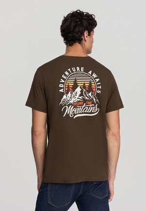 T-shirt in cotone marrone con stampa frontale di montagne, alberi e il testo "Adventure Awaits" in bianco e giallo, con un design vintage.